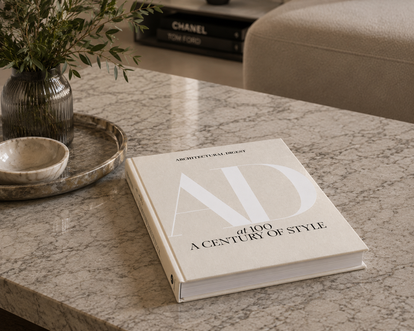A copy of the book 'AD at 100: A Century of Style' by Annette Adjeigui lies open on a light gray marble table, styled with a small ceramic bowl, a textured vase with greenery, and a sofa in the background.