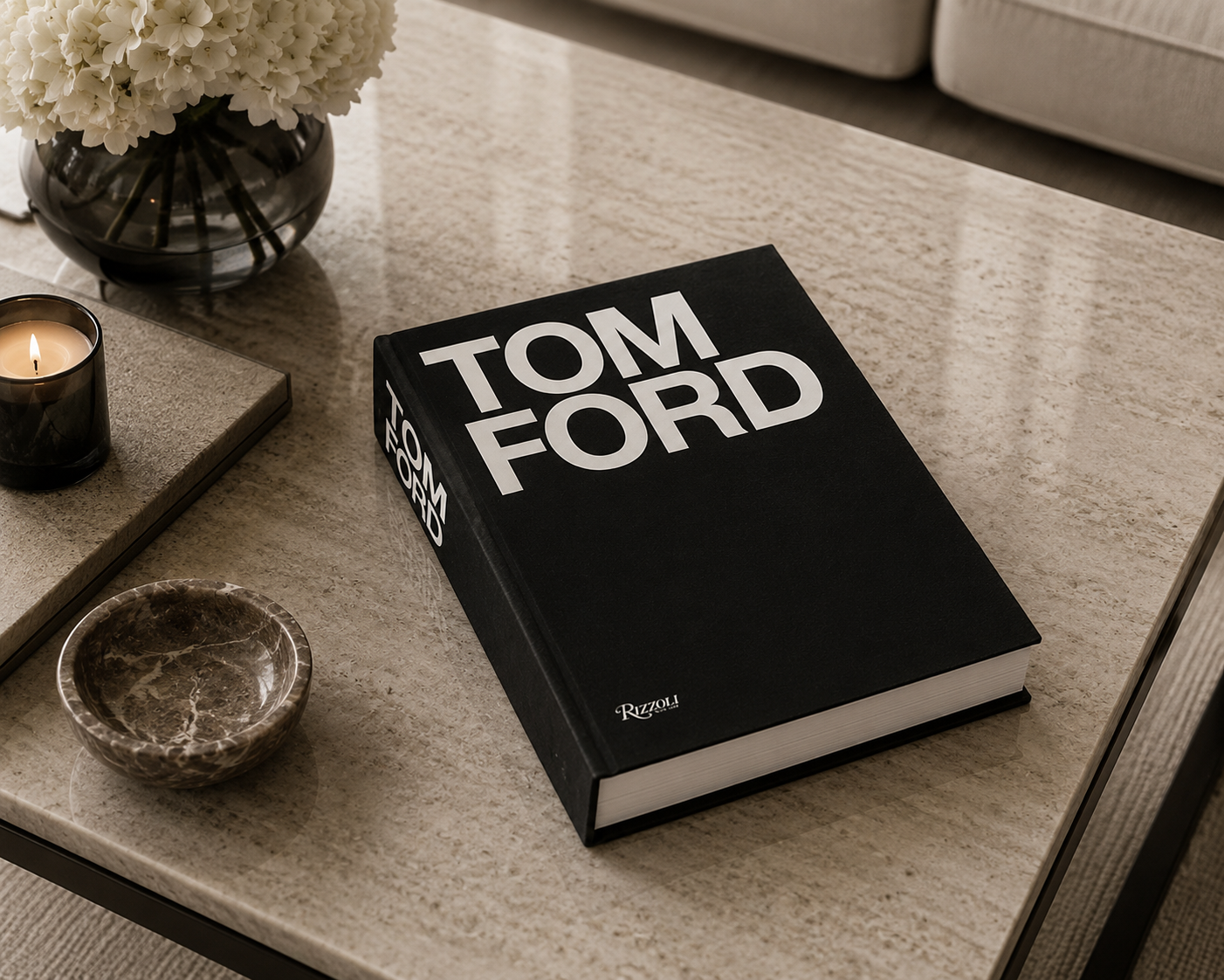 A black hardcover book with bold white 'TOM FORD' lettering on the cover, placed on a light stone tabletop in a styled interior setting with a candle, flowers, and a small bowl.