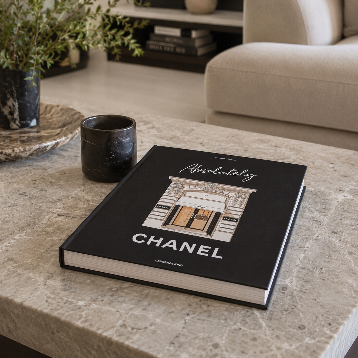 A hardcover book titled 'Absolutely Chanel' by Laurent Mott, placed on a light marble table in a modern living room setting next to a dark ceramic cup.