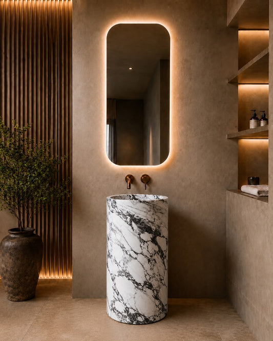 A modern bathroom interior featuring a cylindrical pedestal sink made of white marble with black veining, positioned beneath a vertically oriented illuminated mirror with warm backlighting.